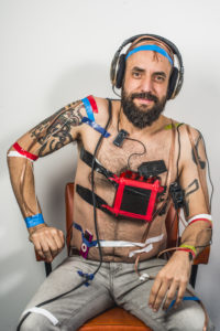 Jonny smiling, topless wearing headphones with sound equipment taped to his chest