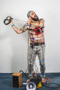 Jonny pulling off headphoes that are taped to his skin. Speakers and megaphone at his feet