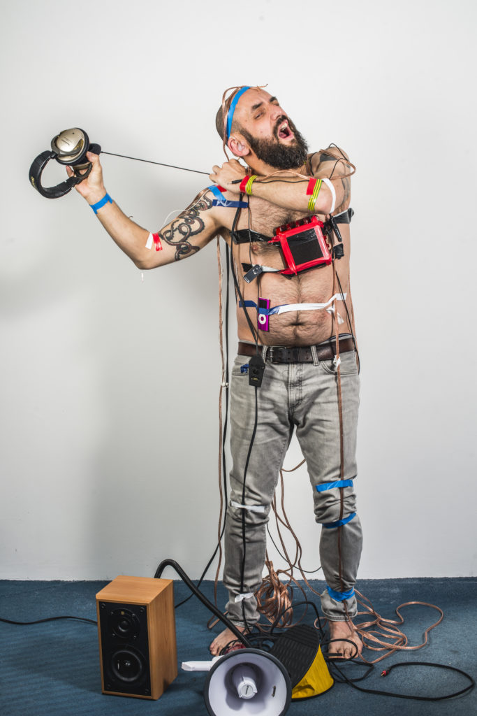 Jonny pulling off headphones that are taped to his skin. Speakers and megaphone at his feet